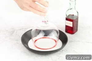 Dipping a gin glass coated in grenadine into caster sugar in a black bowl to make a sugar rim glass with a green bottle of grenadine in the background