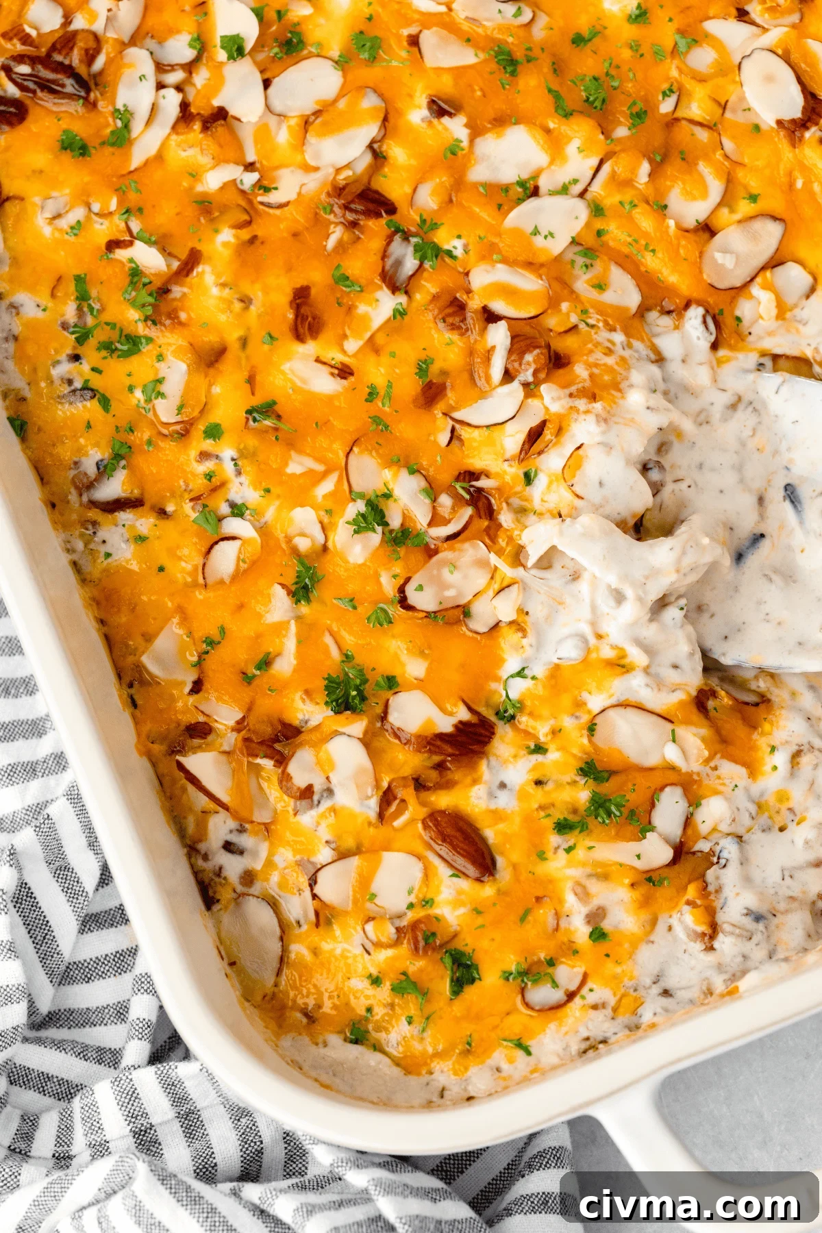 Casserole dish filled with a cheesy chicken casserole, topped with sliced almonds and fresh parsley. The dish is partially scooped out, revealing the creamy interior. A striped cloth is placed beside the dish.