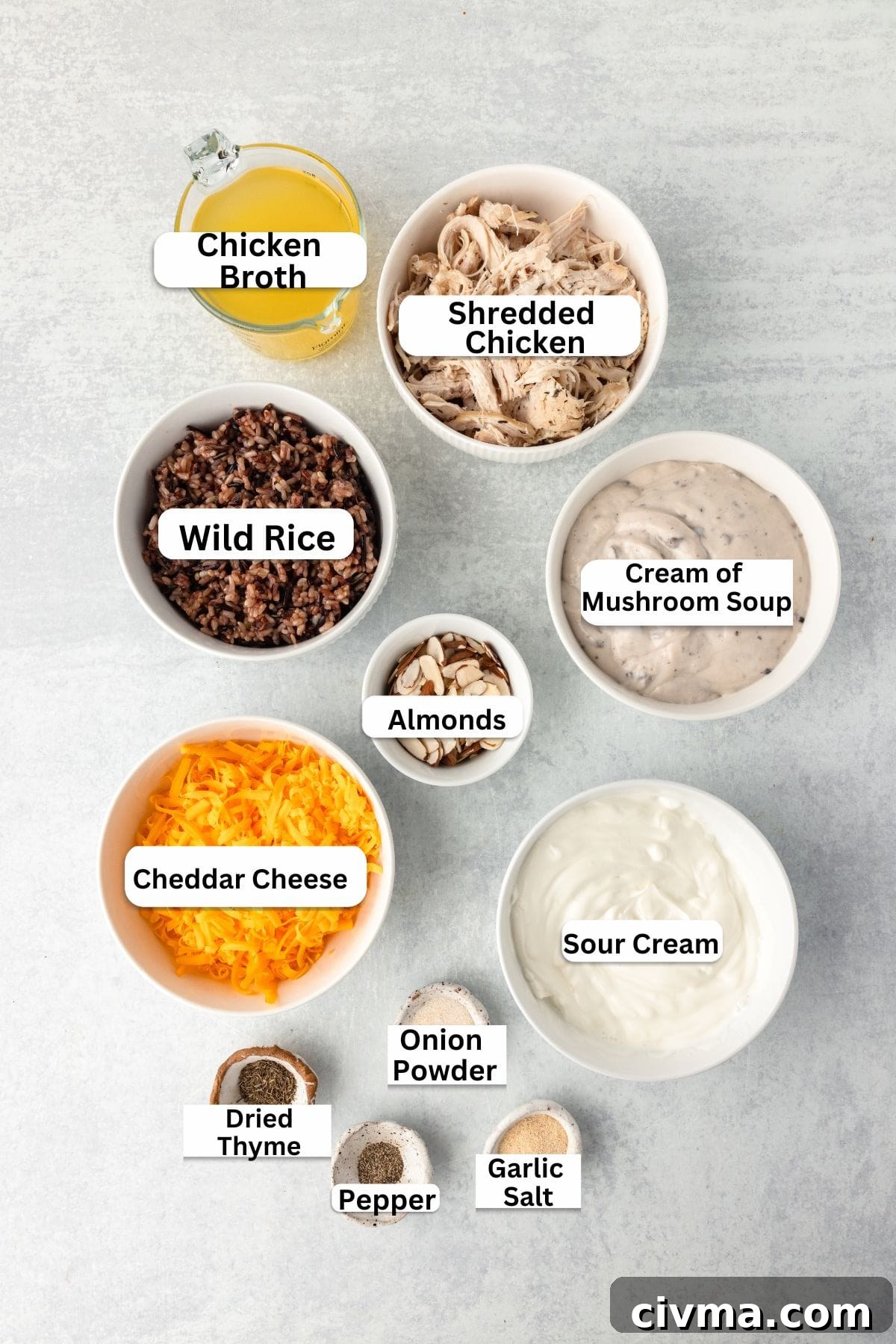 A top-down view of nine labeled bowls containing ingredients for a recipe: chicken broth, shredded chicken, wild rice, cream of mushroom soup, almonds, cheddar cheese, sour cream, dried thyme, pepper, onion powder, and garlic salt.
