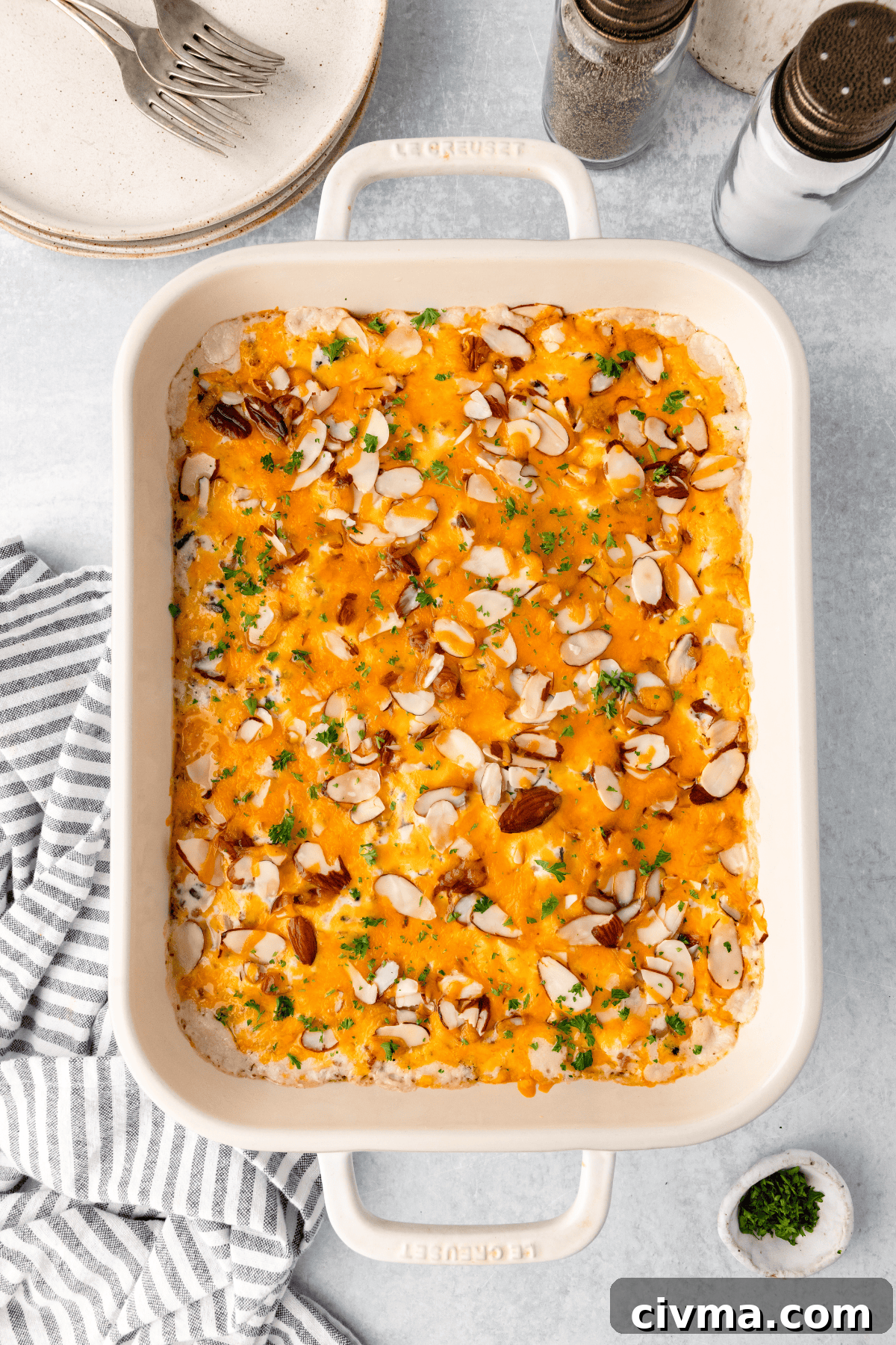 A casserole dish filled with a baked dish topped with melted cheese, sliced almonds, and chopped herbs. The dish is placed on a light-colored surface next to a striped cloth, with plates and salt and pepper shakers in the background.