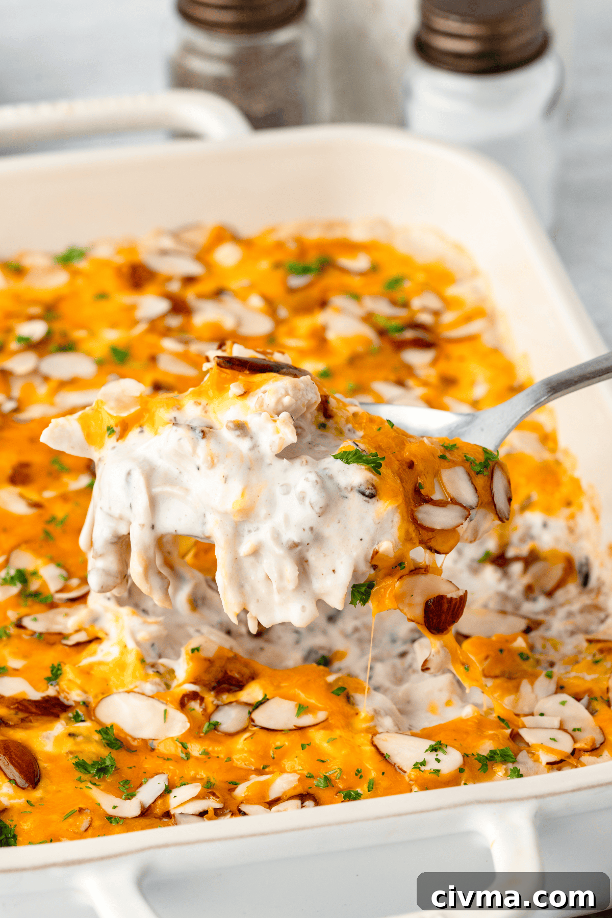 A close-up of a creamy baked casserole being scooped from a dish. It has a golden cheese topping, sprinkled with sliced almonds and fresh parsley. Two salt and pepper shakers are blurred in the background.