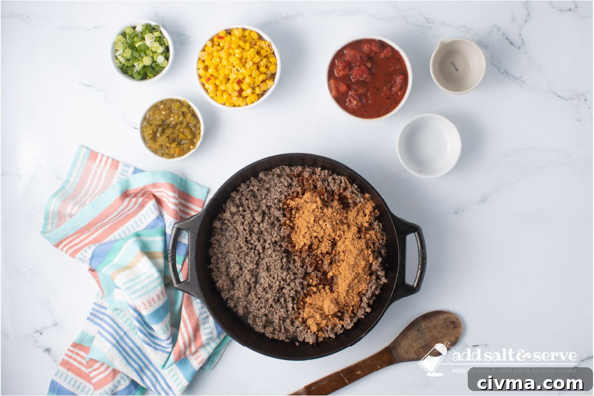 Cast iron skillet with cooked ground beef and taco seasoning surrounded by bowls with mexi-corn, green chilis, fire-roasted diced tomatoes, water, and chopped green onions and a wooden spoon.
