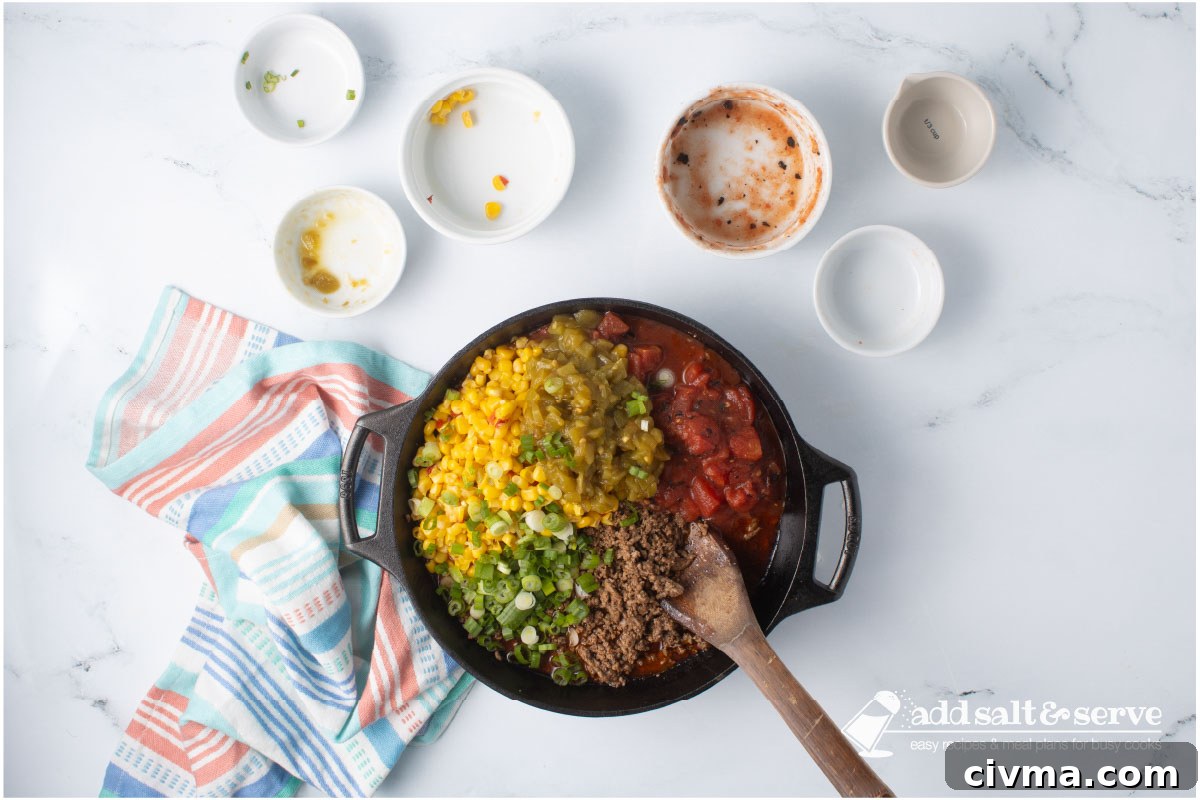Cast iron skillet with seasoned cooked ground beef, mexi-corn, green chilis, fire-roasted diced tomatoes, and chopped green onions with a wooden spoon.