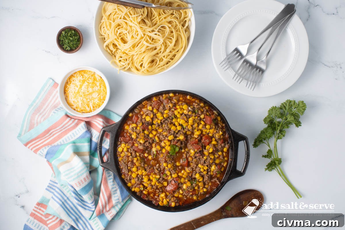 Cast iron skillet of Mexican Spaghetti sauce, a dish of shredded cheddar cheese, and a bowl of cooked spaghetti on a counter next to fresh parsley.