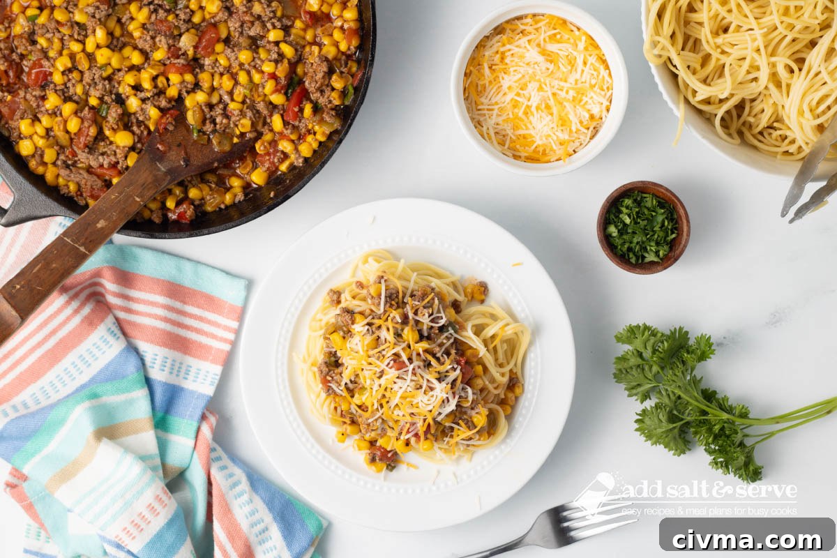 Plate of Mexican Spaghetti topped with shredded cheese beside a bowl of cooked spaghetti, a pot of Mexican Spaghetti sauce, and a dish of shredded cheddar cheese on a counter next to fresh tomatoes and parsley.