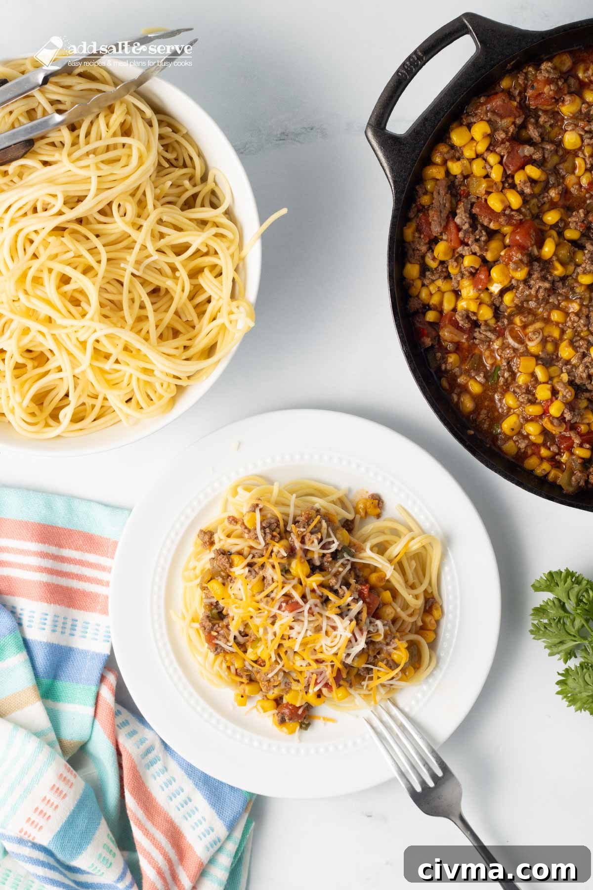 Plate of Mexican Spaghetti topped with shredded cheese on a counter beside a bowl of cooked spaghetti and a pot of Mexican Spaghetti sauce.