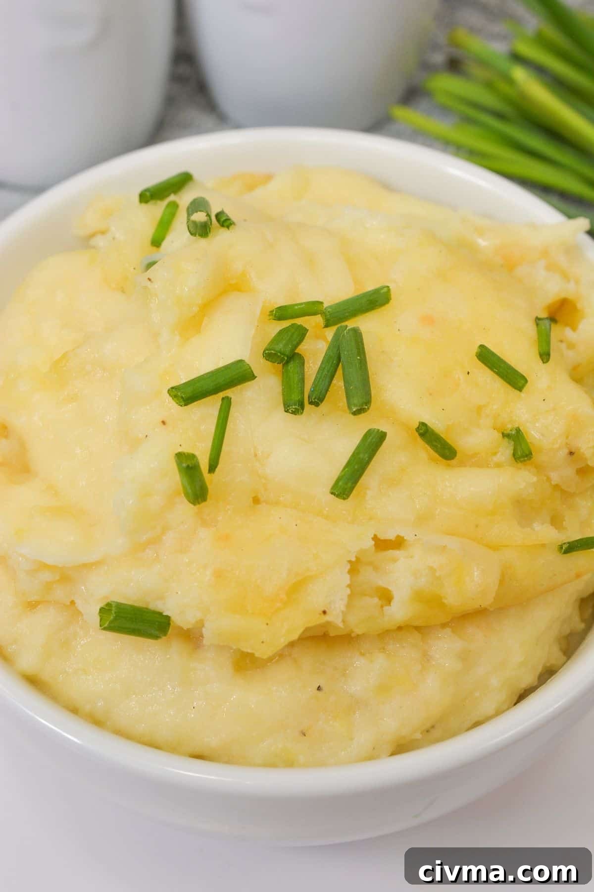 A white bowl filled with cheesy mashed potatoes, garnished with chopped chives, placed on a white surface.
