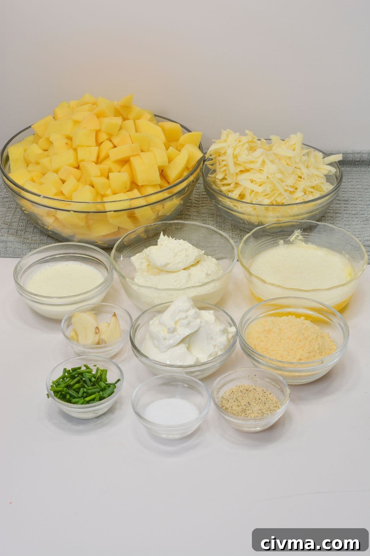Assorted glass bowls filled with diced potatoes, shredded cheese, sour cream, cream cheese, butter, garlic, seasonings, and chopped chivesare arranged on a white surface.