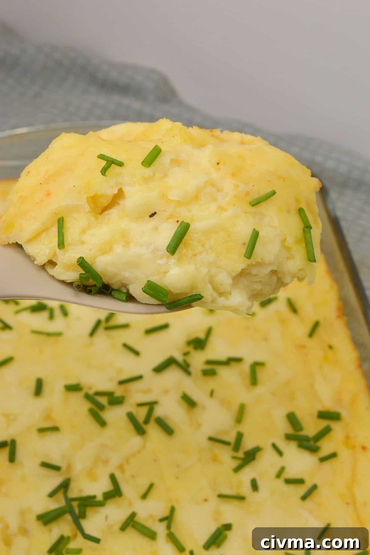 A serving of Cheesy Mashed Potatoes casserole with chopped chives on top is lifted from a baking dish.