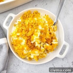 A round white dish filled with creamy Cheesy Party Potatoes casserole, topped with golden, crispy cornflakes, with a spoon resting inside the dish.