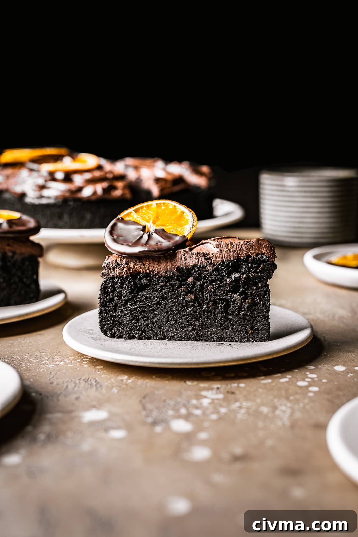 A slice of cake with thick chocolate frosting on a plate. A chocolate dipped dried orange slice sits on top.