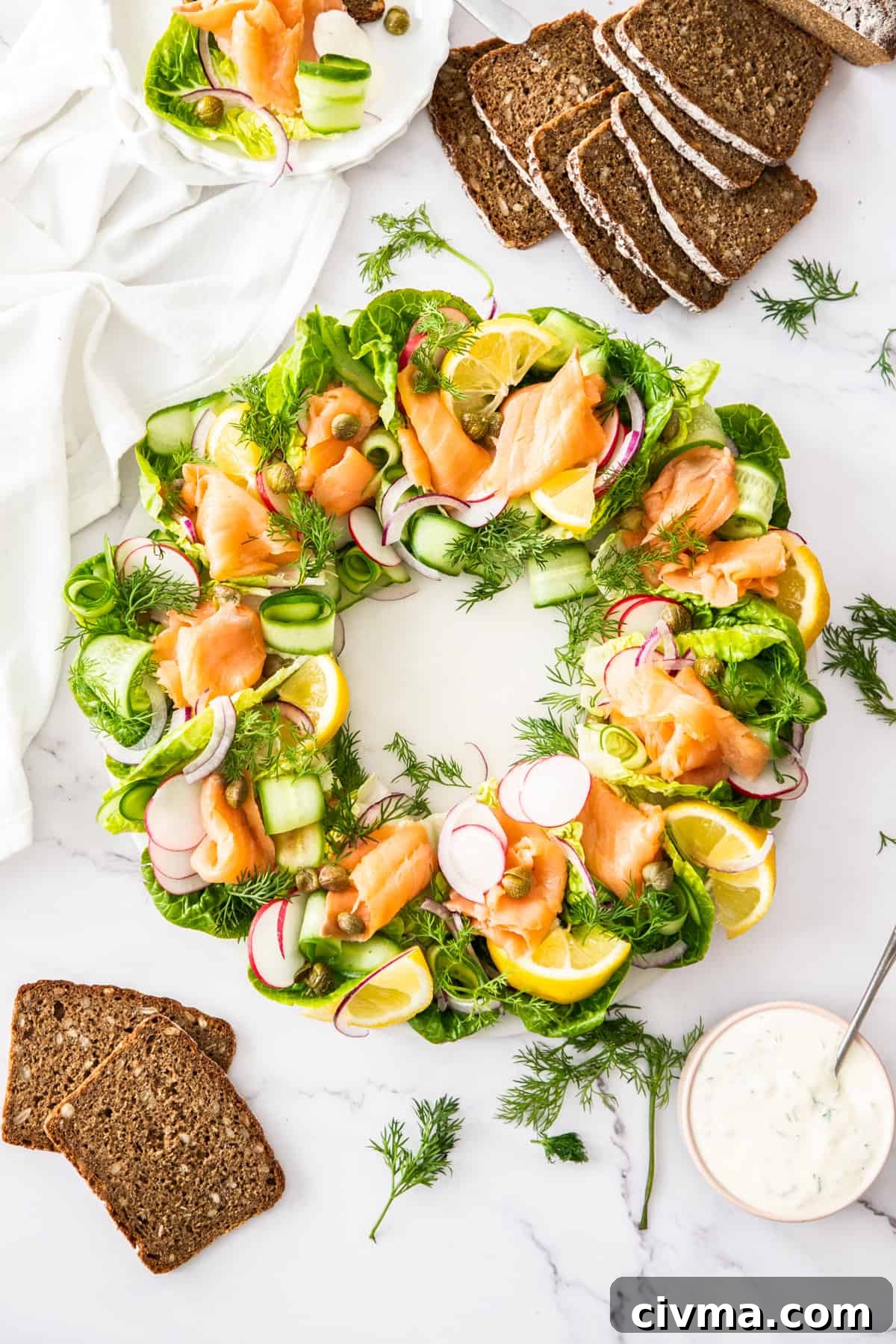 Smoked Salmon Wreath on a round white plate, sitting on a marble bench with some dressing and slices of rye bread around edge.